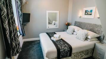 Un dormitorio ordenado con cama, mesitas de noche, un espejo y un televisor montado en la pared.