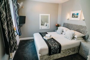 Un dormitorio ordenado con cama, mesitas de noche, un espejo y un televisor montado en la pared.