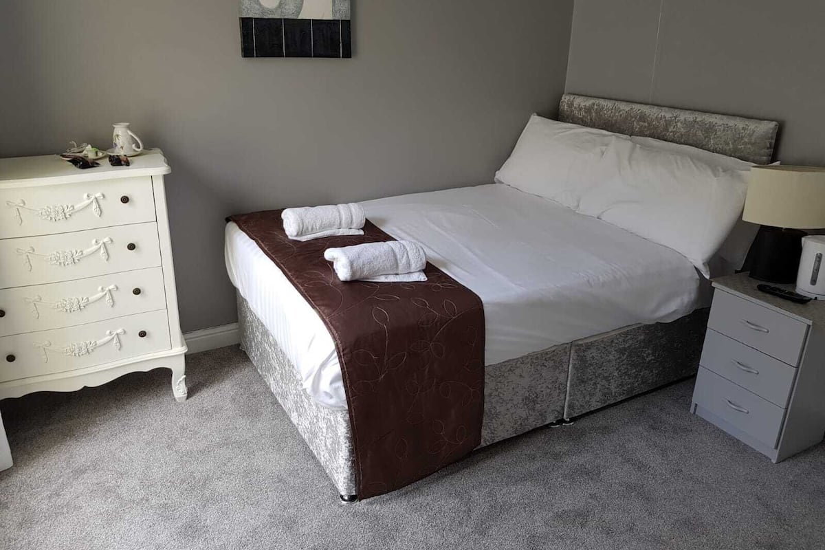 A neatly made bed with a brown bedspread, white pillows, and two folded towels. A white dresser with drawers is to the left, and a small bedside table with a lamp is to the right.