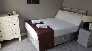 A neatly made bed with a brown bedspread, white pillows, and two folded towels. A white dresser with drawers is to the left, and a small bedside table with a lamp is to the right.