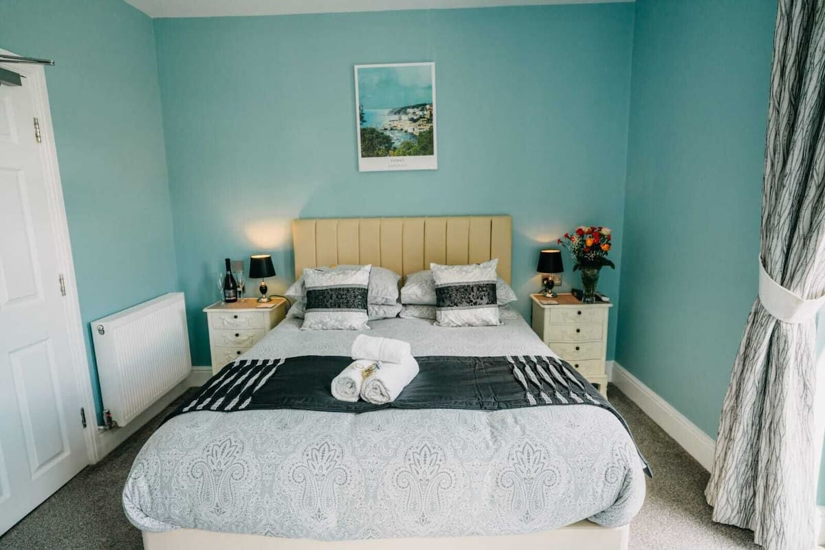 A bedroom with a bed, two bedside tables, a nightstand, a radiator, a picture on the wall, and a curtain.