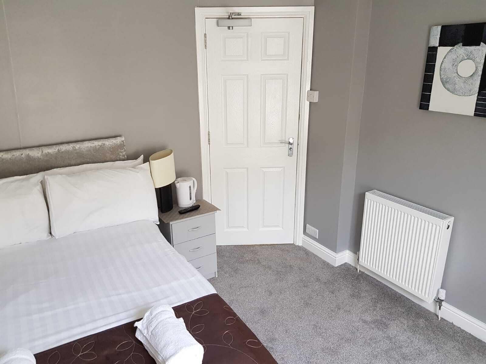A neatly made bed with white linens, a bedside table with a lamp and a kettle, a white door, and a radiator on the wall.