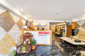A hotel reception area with a reception desk, a wall-mounted security camera, a sofa, a refrigerator, and a decorative altar with offerings.