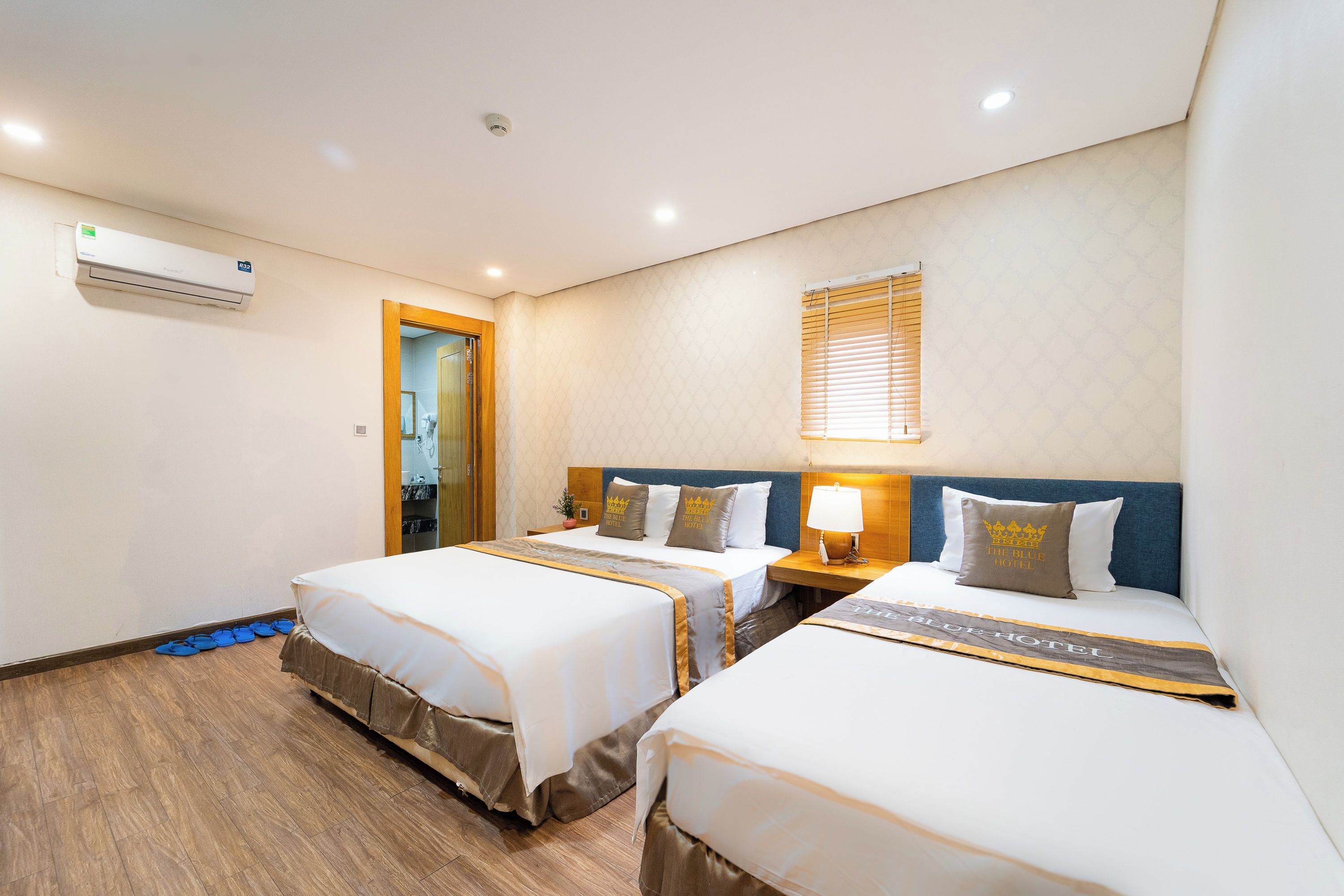 A hotel room with two beds, a bedside table with a lamp, a window with blinds, and an air conditioning unit on the wall.