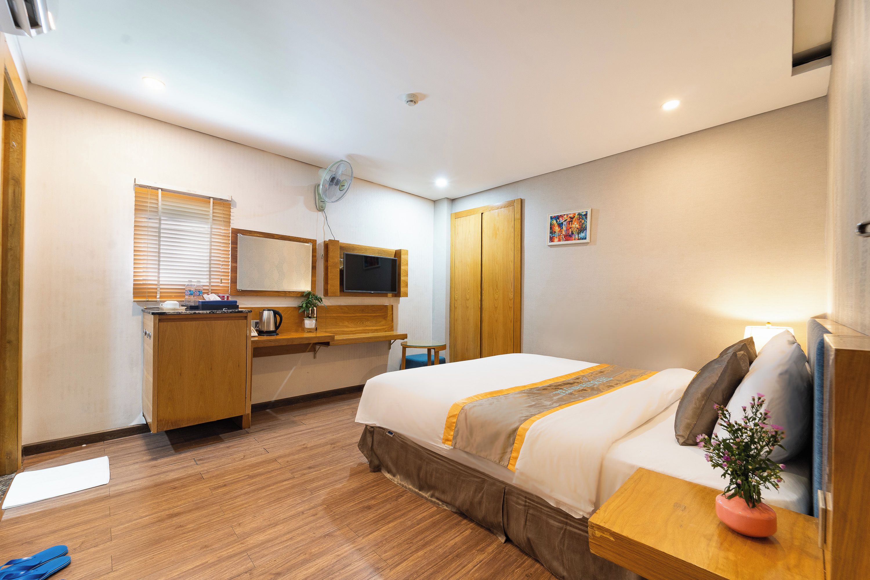 Une chambre d’hôtel avec un lit, un bureau, une télévision, une fenêtre avec des stores et un petit tableau accroché au mur.