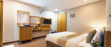 A hotel room with a bed, a desk, a television, a window with blinds, and a small painting on the wall.