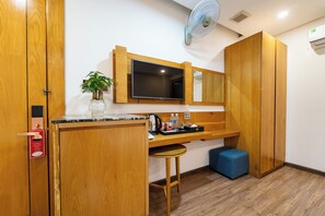 Una habitación compacta con un escritorio de madera, un televisor montado en la pared, un taburete pequeño, un reposapiés azul y una planta en maceta.