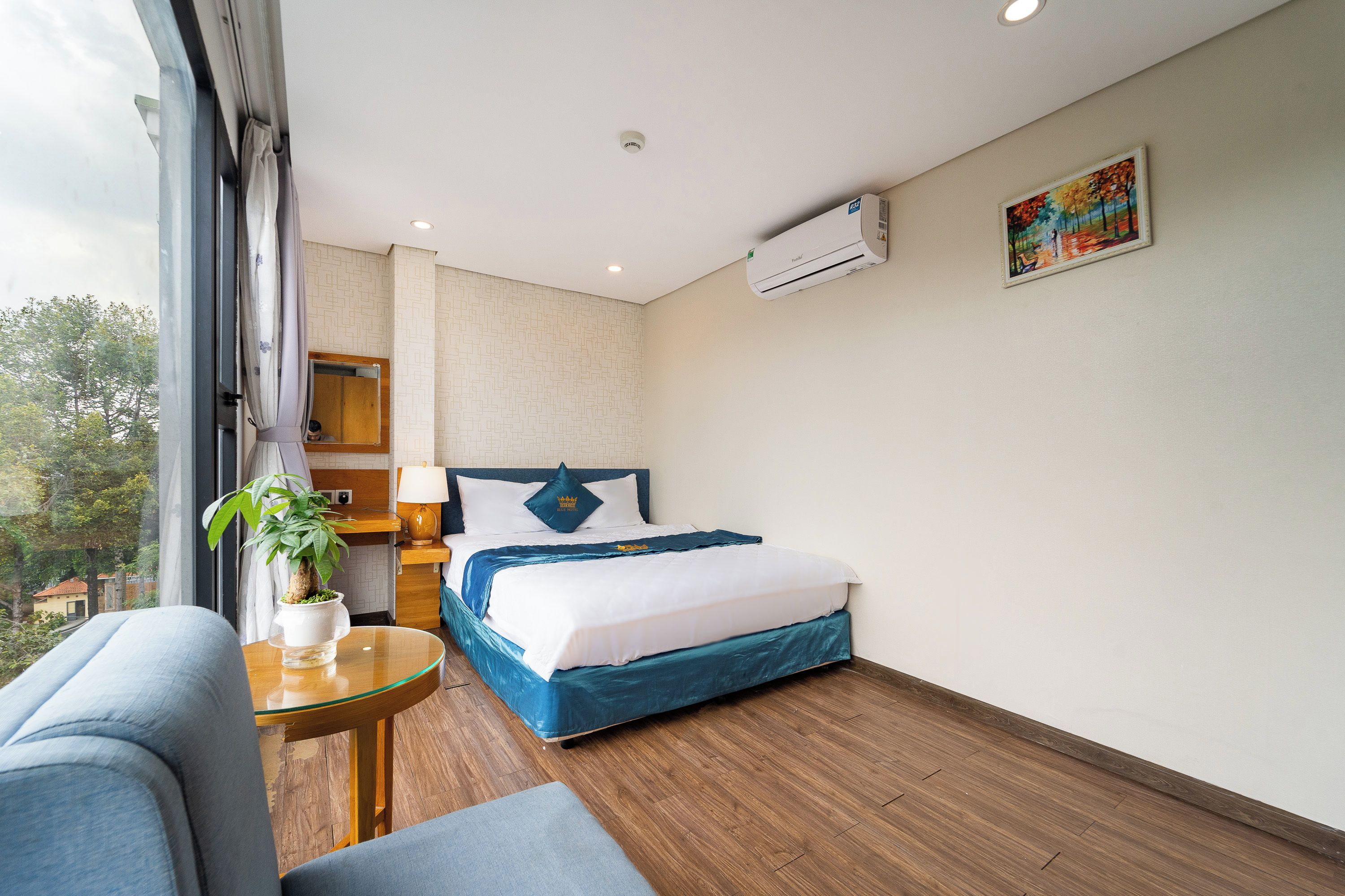 Habitación de hotel con cama, mesita de noche, planta en maceta, ventana y aire acondicionado.