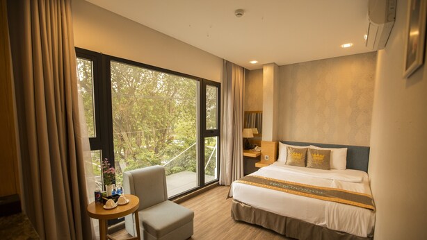 Quarto de hotel com uma janela grande, uma cama com cabeceira decorativa, uma pequena mesa com uma cadeira e uma mesa de cabeceira com um abajur.
