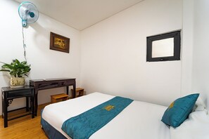 Habitación de hotel con cama, escritorio, silla, planta en maceta, ventilador de pared, cuadro enmarco y espejo.