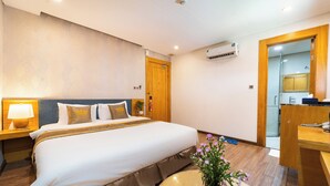 A hotel room with a large bed, a bedside table with a lamp, a wall-mounted air conditioner, and a wooden door leading to a bathroom. - The Blue Hotel (Ho Chi Minh City)