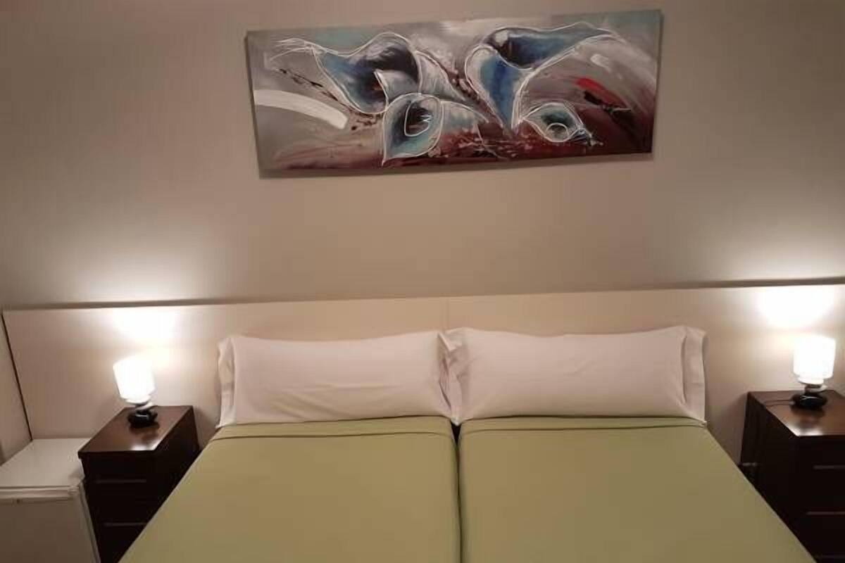 Una cama doble con una colcha verde, dos mesitas de noche con lámparas, y un cuadro en la pared.