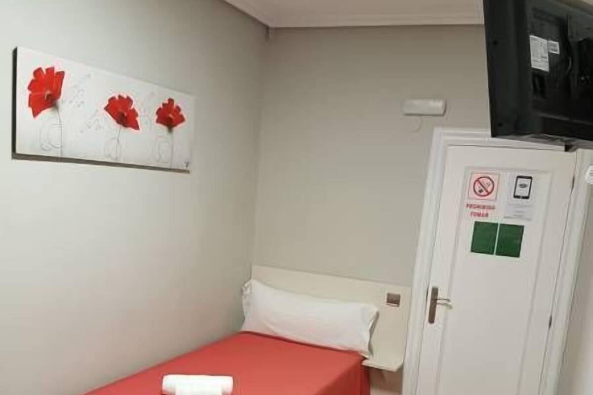 Una habitación pequeña y limpia con una cama individual, una mesa y un mando a distancia, además de una puerta con un letrero de 'no fumar'.