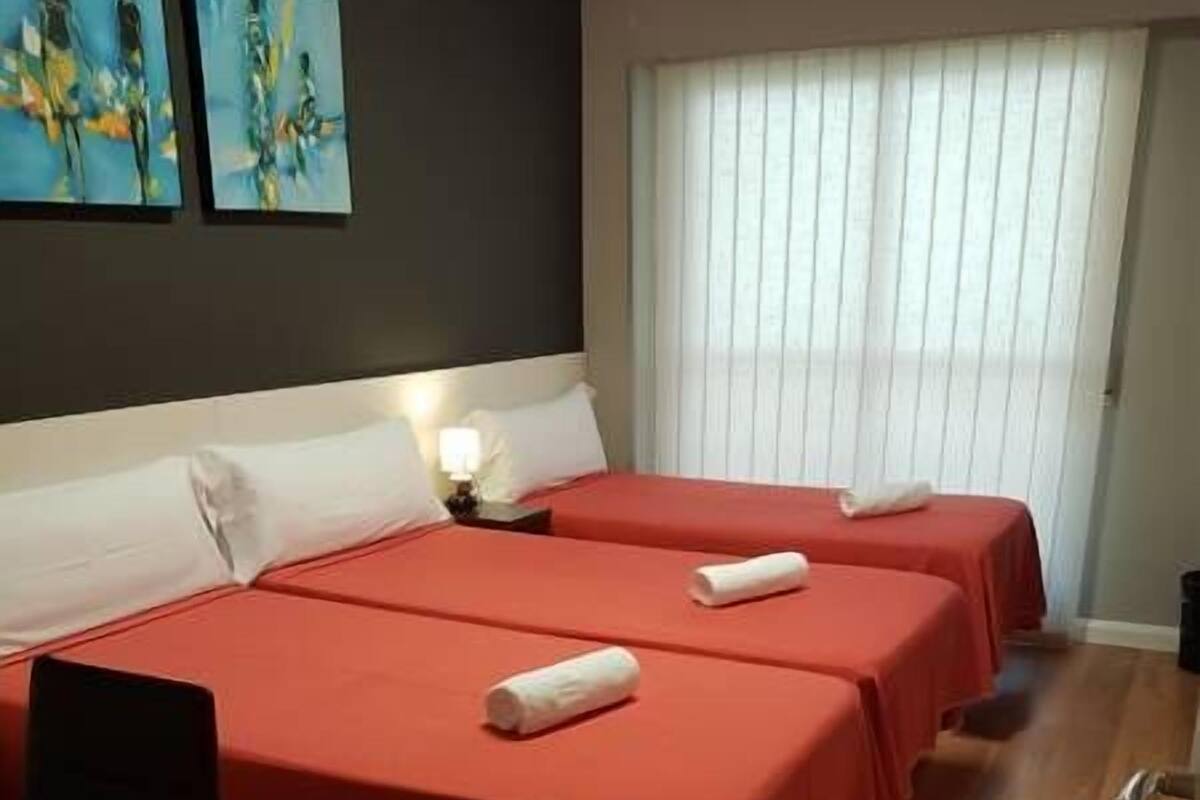 Habitación de hotel con dos camas, un escritorio y una silla. Hay cuadros en la pared y una ventana con persianas.