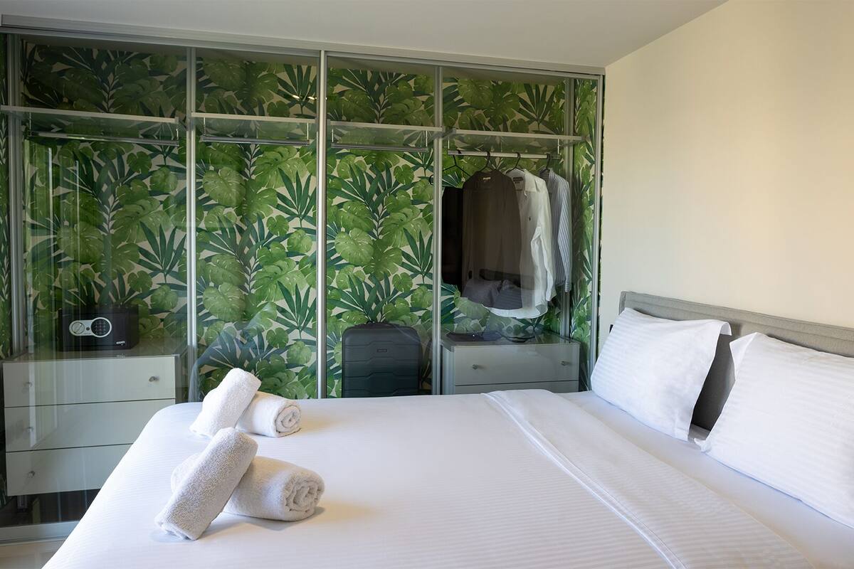 A hotel room with a bed, a nightstand, a wardrobe with a tropical leaf pattern, and a suitcase.