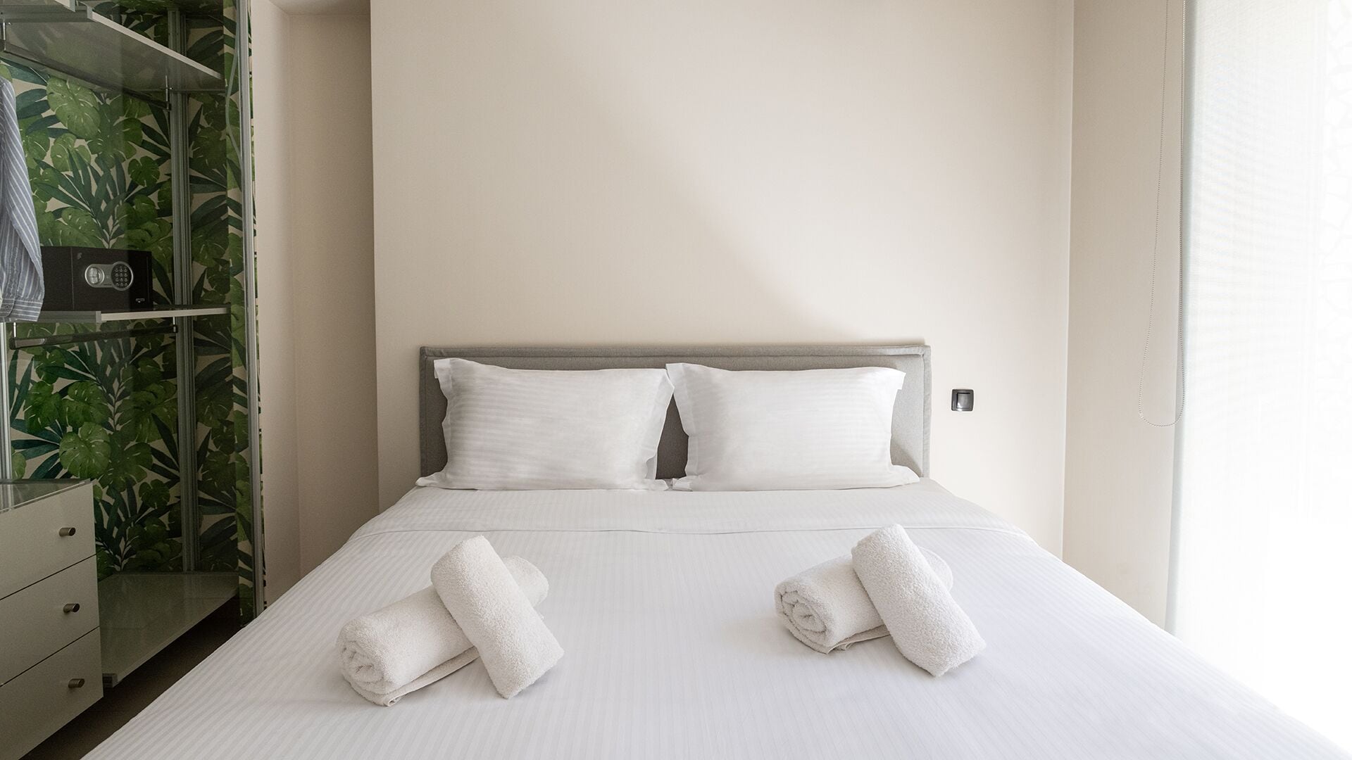 A neatly made bed with two pillows and two rolled towels, a bedside table, and a wardrobe with a plant.