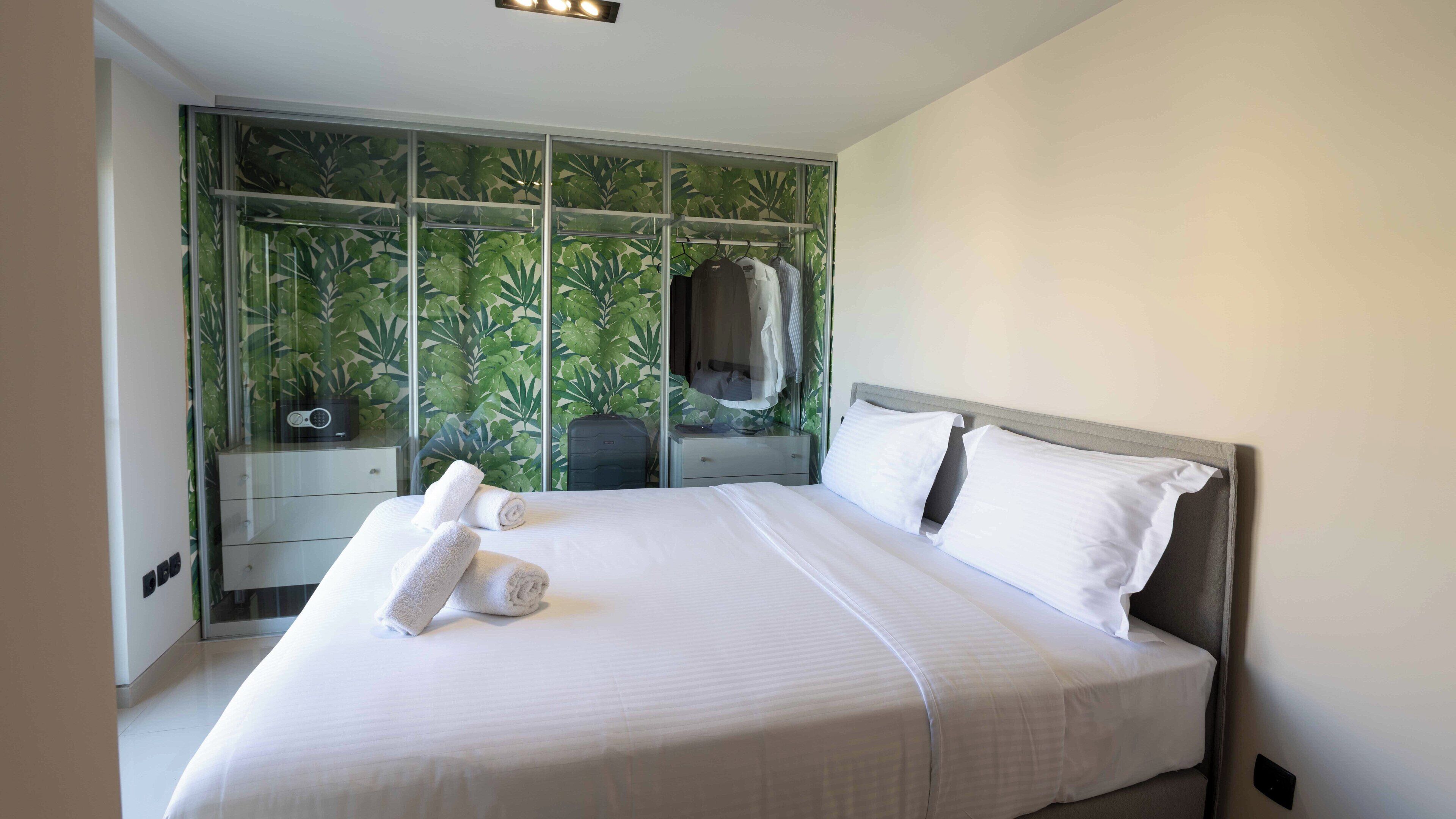 A neatly made bed with white linens and two rolled towels, a bedside table with a lamp, and a wardrobe with a green leafy wallpaper backdrop.