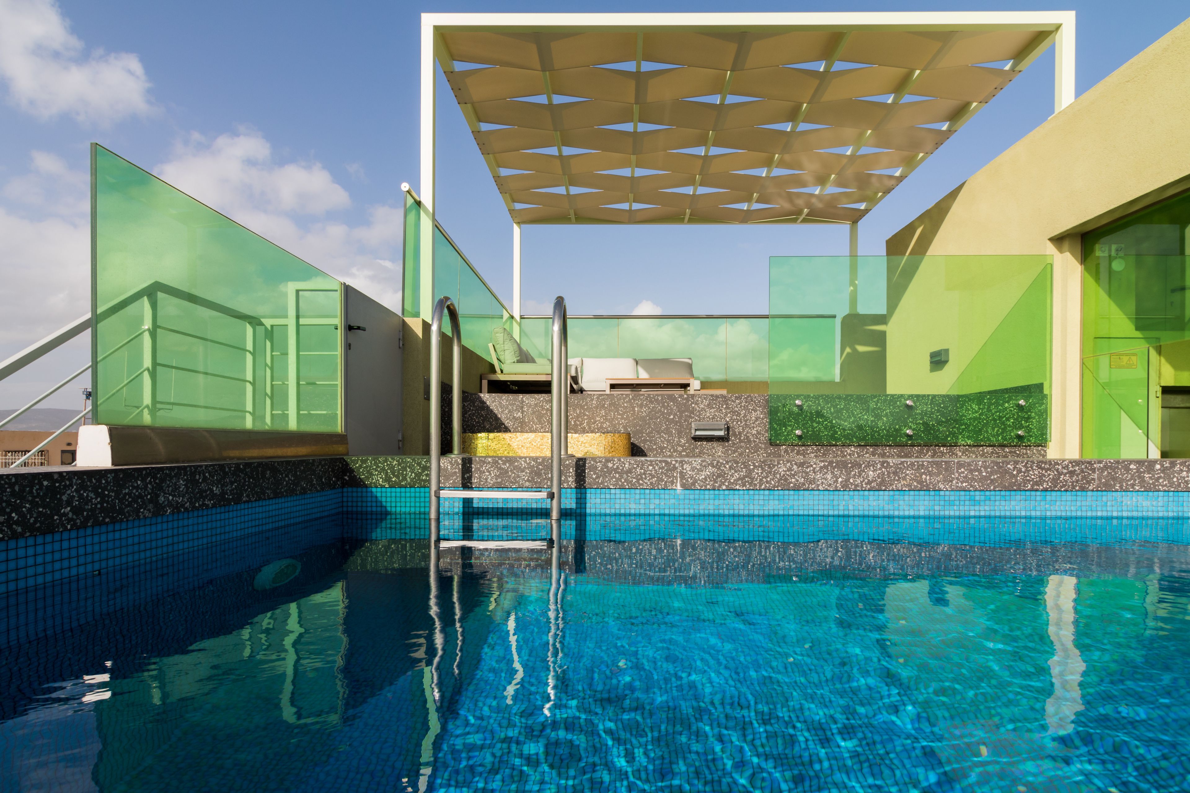 Un área de piscina moderna con agua azul cristalina, cerramiento de vidrio y una zona de estar con sombra.