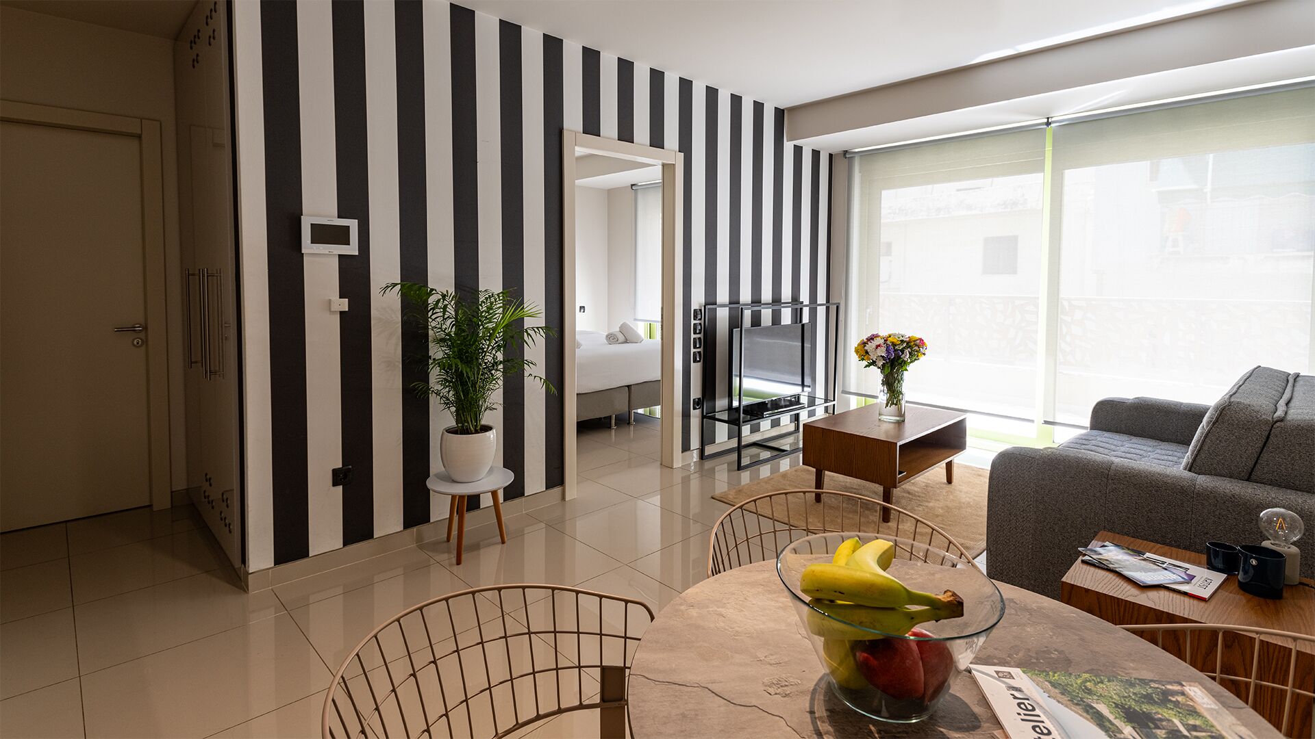 A modern hotel room with a striped wall, a bed, a TV, a dining area with a table and chairs, and a sofa.