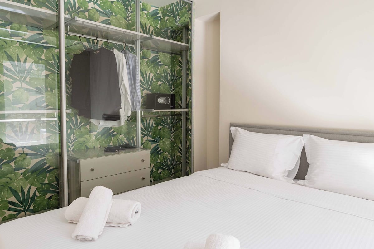 A hotel room with a bed, white pillows, a bedside table, a mirror, and a wardrobe with a tropical leaf pattern.