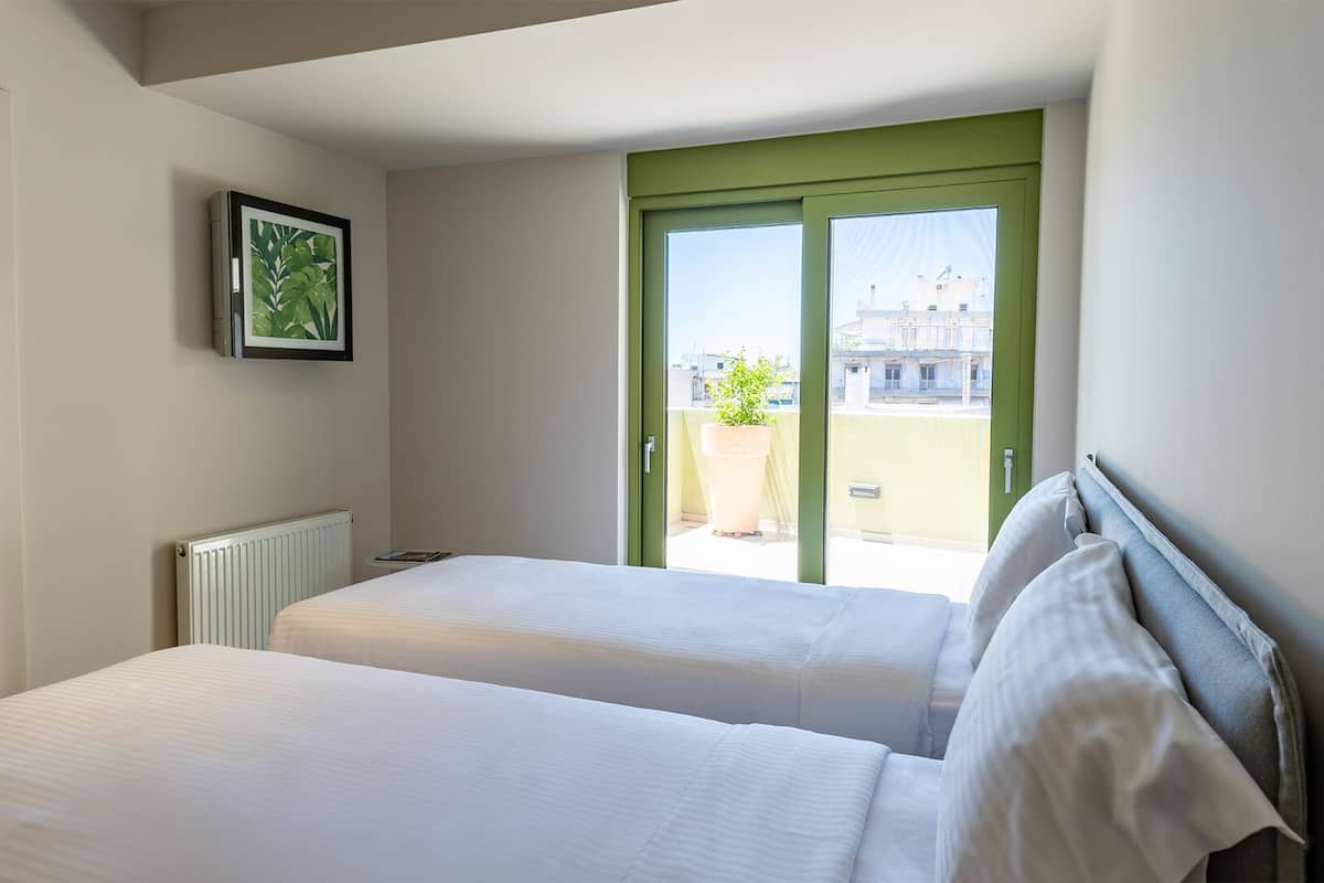 A hotel room with two beds, a radiator, a framed picture, and a view of a balcony with a potted plant.