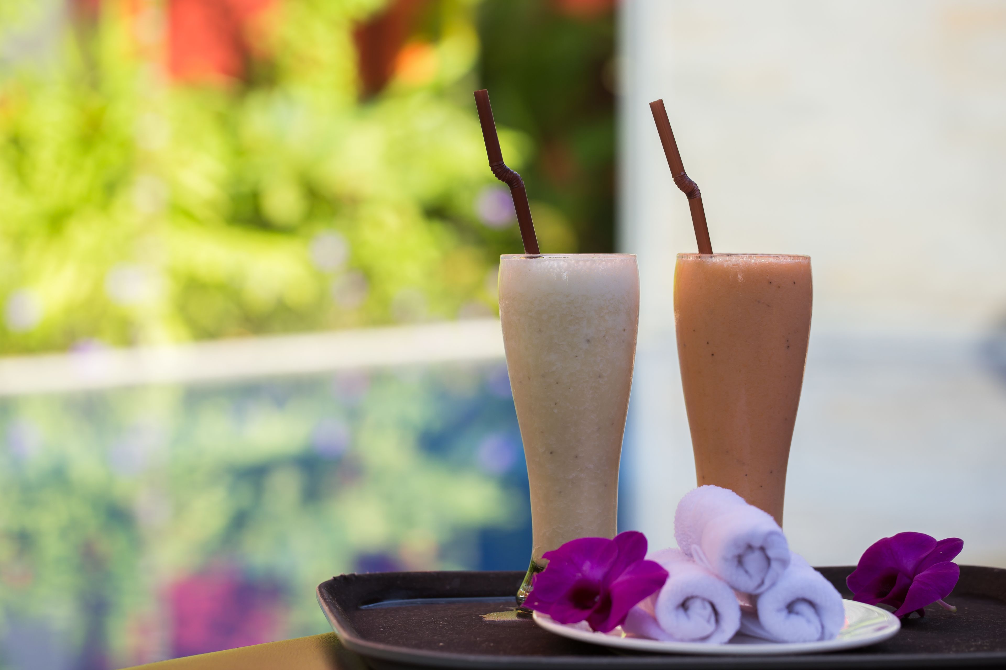 Dos vasos altos de una bebida con pajitas, un plato con toallas enrolladas y flores moradas en una bandeja junto a una piscina.