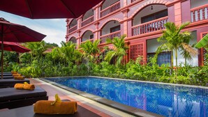 Un espace aménagé au bord de la piscine, avec des chaises longues et des parasols, entouré d’une végétation luxuriante et d’un grand bâtiment doté de balcons.