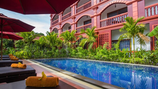A poolside area with lounge chairs and umbrellas, surrounded by lush greenery and a large building with balconies. - Bou Savy Villa (Siem Reap)