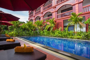 A poolside area with lounge chairs and umbrellas, surrounded by lush greenery and a large building with balconies. - Bou Savy Villa (Siem Reap)