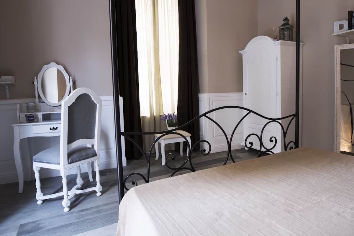 A bedroom with a bed, a dresser, a vanity with a mirror, a chair, and a small table with a vase of flowers.