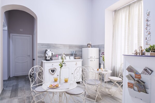 A modern kitchen with white cabinetry, a dining area with metal chairs, and a small table with a fruit plate. - Dimora Partenopea (Naples)