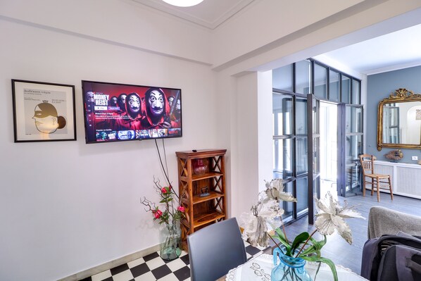 Uma sala de estar moderna com uma televisão de tela plana grande, uma estante de madeira, uma mesa de jantar com cadeiras e uma grande janela com vista para a cidade.