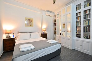 A neatly arranged bedroom with a large bed, bedside lamps, a bookshelf, and an air conditioning unit.