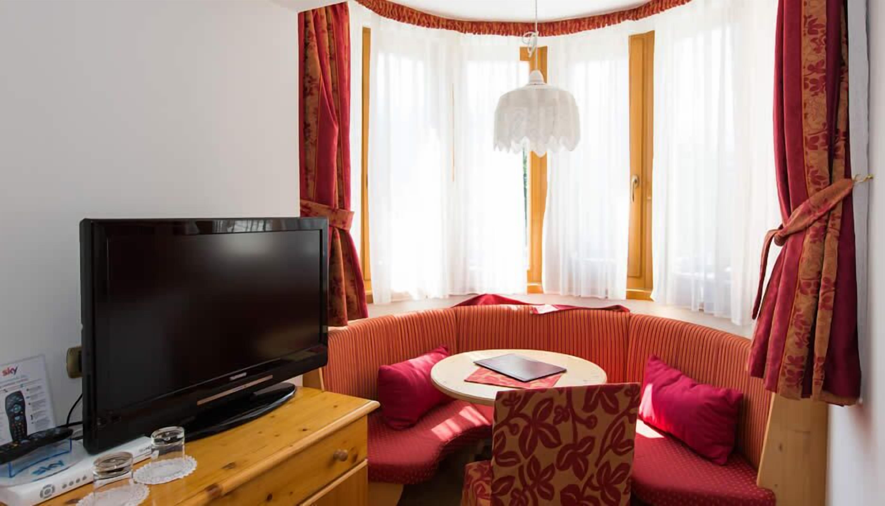 A cozy room with a large flat-screen TV on a wooden dresser, a round table with a laptop, and a comfortable seating area with a sofa and pillows.