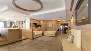 A modern hotel lobby with a reception desk, two sofas, a large mirror, and a wooden accent wall. - Hotel Cristina (Pinzolo)