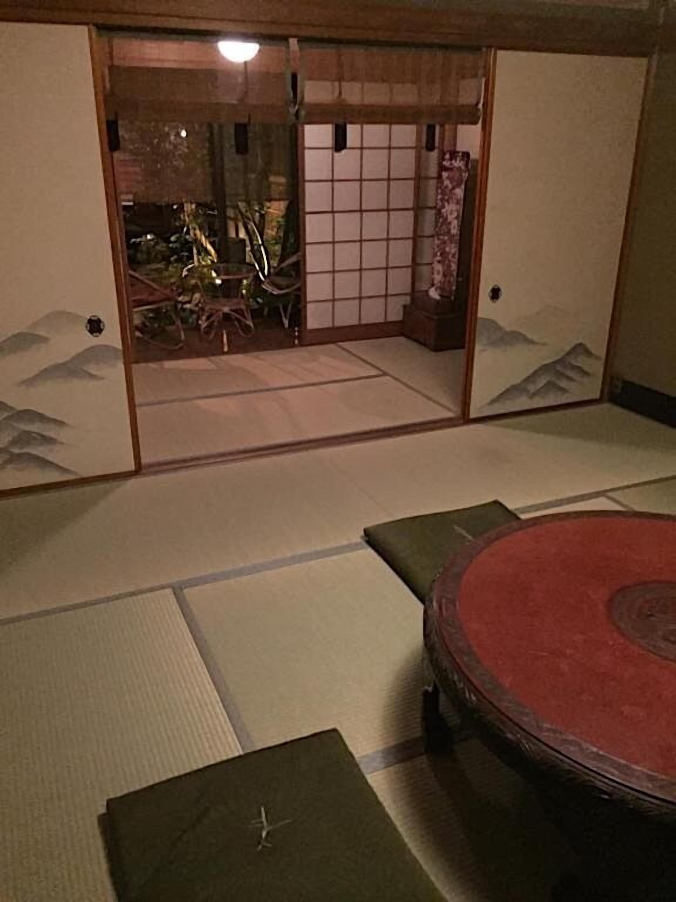 Una habitación con esteras de tatami, una mesa baja, cojines, una puerta corredera que da a un jardín y vistas a montañas.