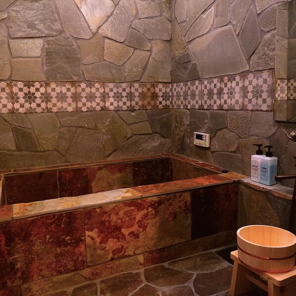 Un baño de paredes de piedra con una bañera, un taburete de madera y un dispensador de jabón.