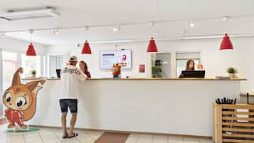 Un área de recepción con mostrador blanco, lámparas colgantes rojas, una pantalla digital y una decoración con un personaje de caricatura.