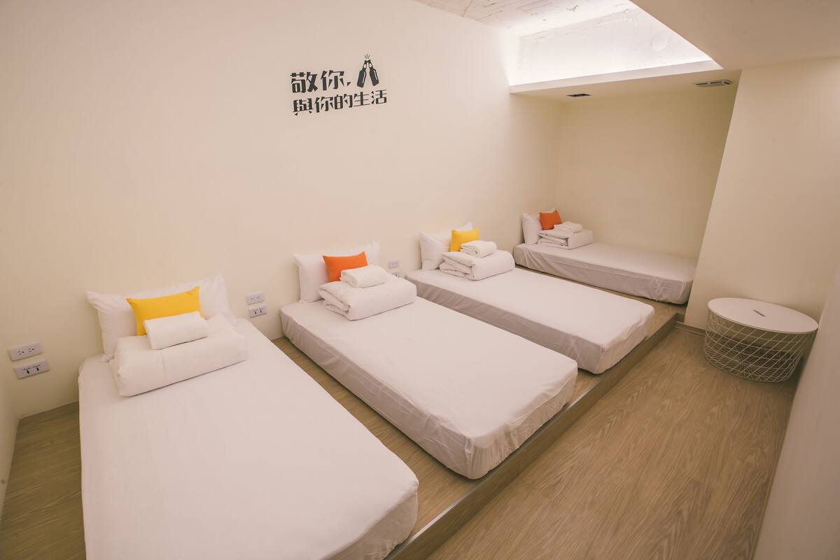 A hotel room with four single beds, each with white bedding and orange pillows. There is a skylight above the beds, and a small trash bin is visible to the right.