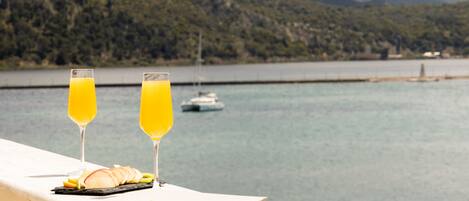 Une table avec un verre de jus d’orange, un verre de vin blanc et une assiette de nourriture, donnant sur une étendue d’eau avec des montagnes en arrière-plan.