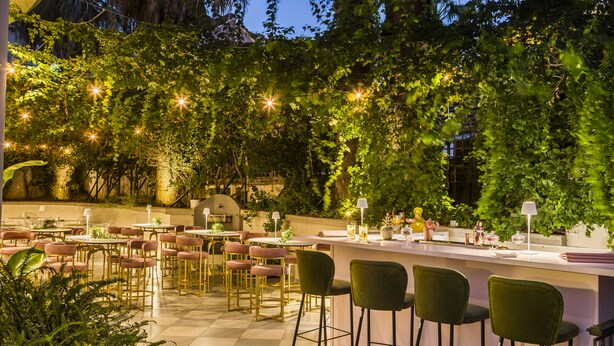 Un espacio exterior para comer con mesas y sillas, rodeado de abundante vegetación e iluminación con luces colgantes.