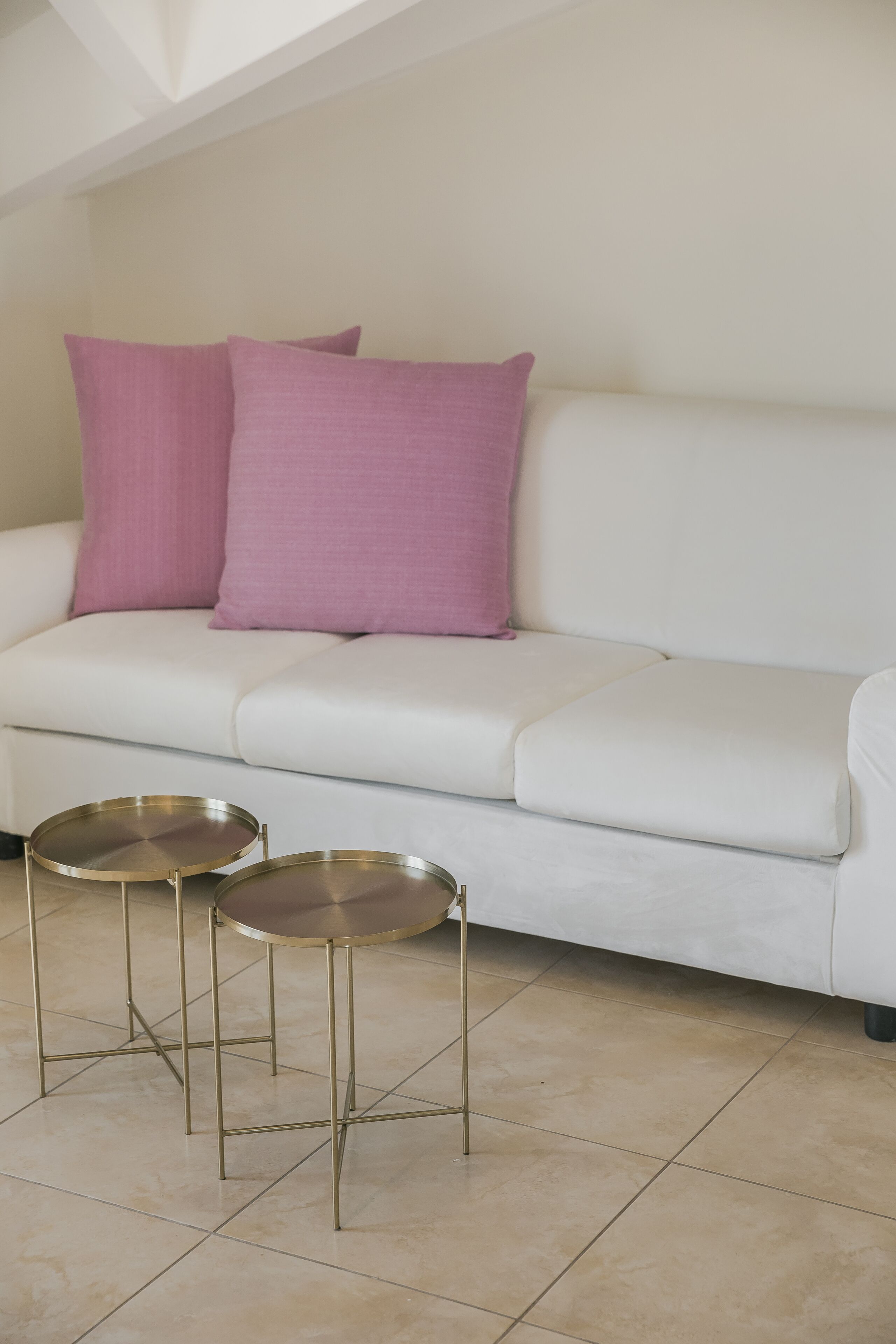 Un sofá blanco con dos mesas redondas de metal delante y dos almohadones morados sobre el sofá.