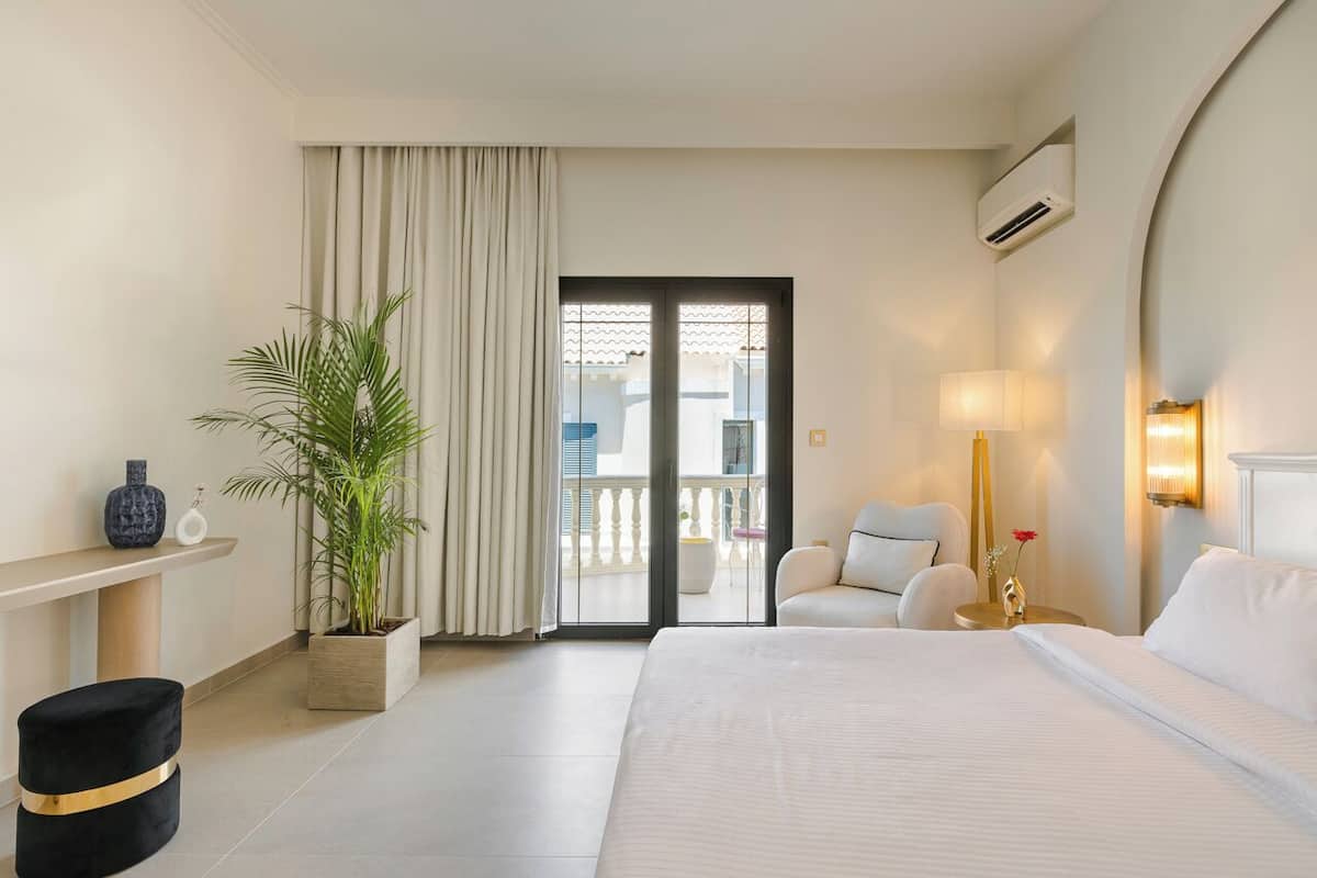 A hotel room with a large bed, a potted plant, a sofa, a small table, and a balcony with a view.