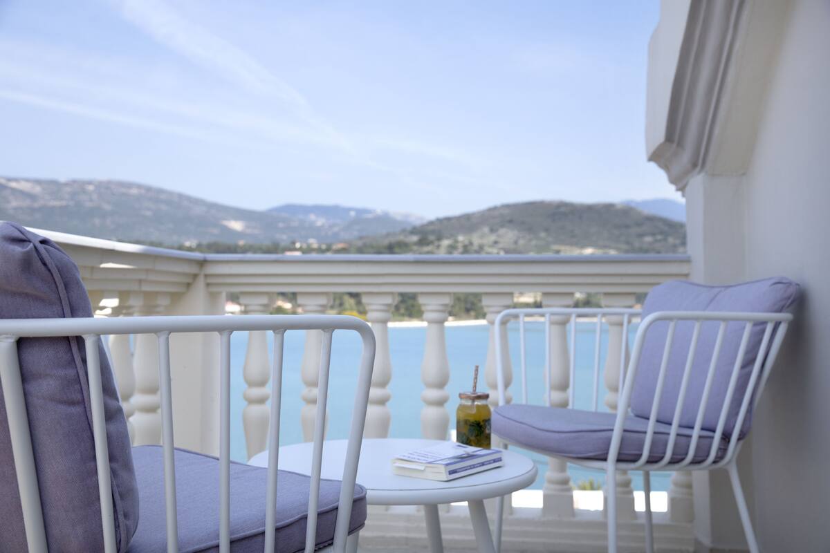 A balcony with two white metal chairs, a small round table, and a view of mountains in the background.