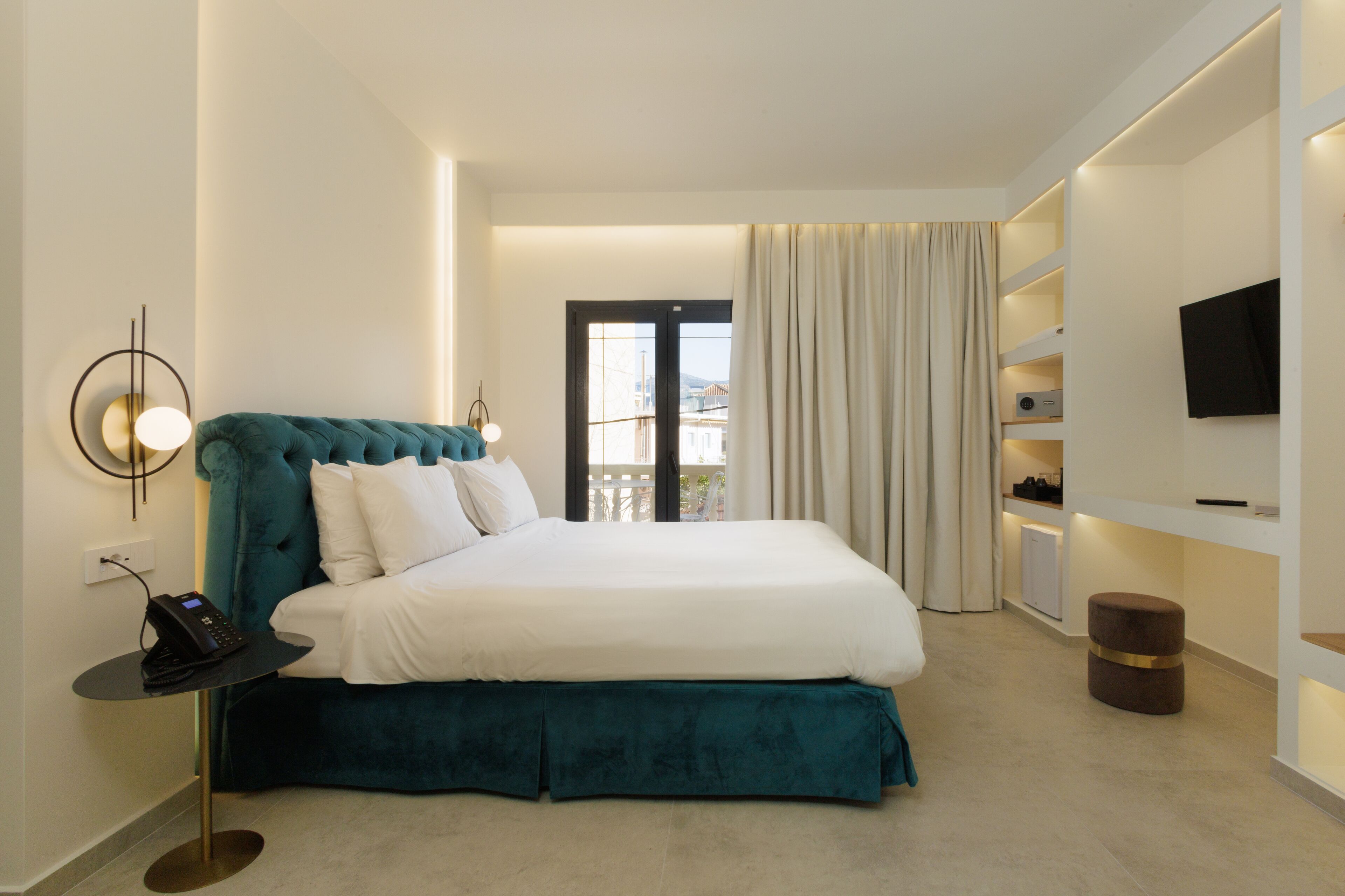 A modern hotel room with a large bed, a bedside table with a phone, a TV on the wall, and a view of the outdoors through a door.