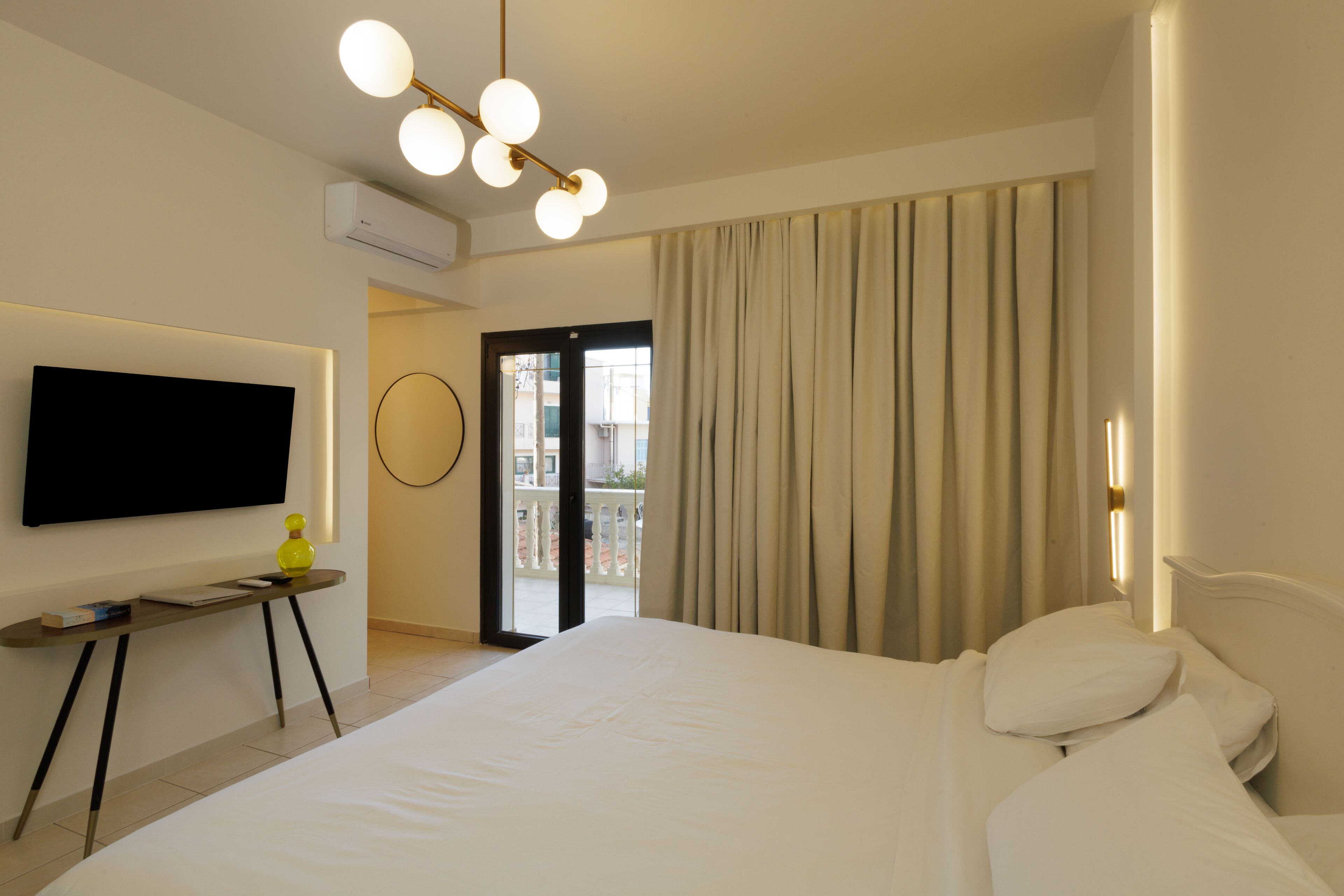 Habitació d'hotel amb un llit, una televisió penjada a la paret, una taula petita amb una làmpada i vista al balcó exterior.