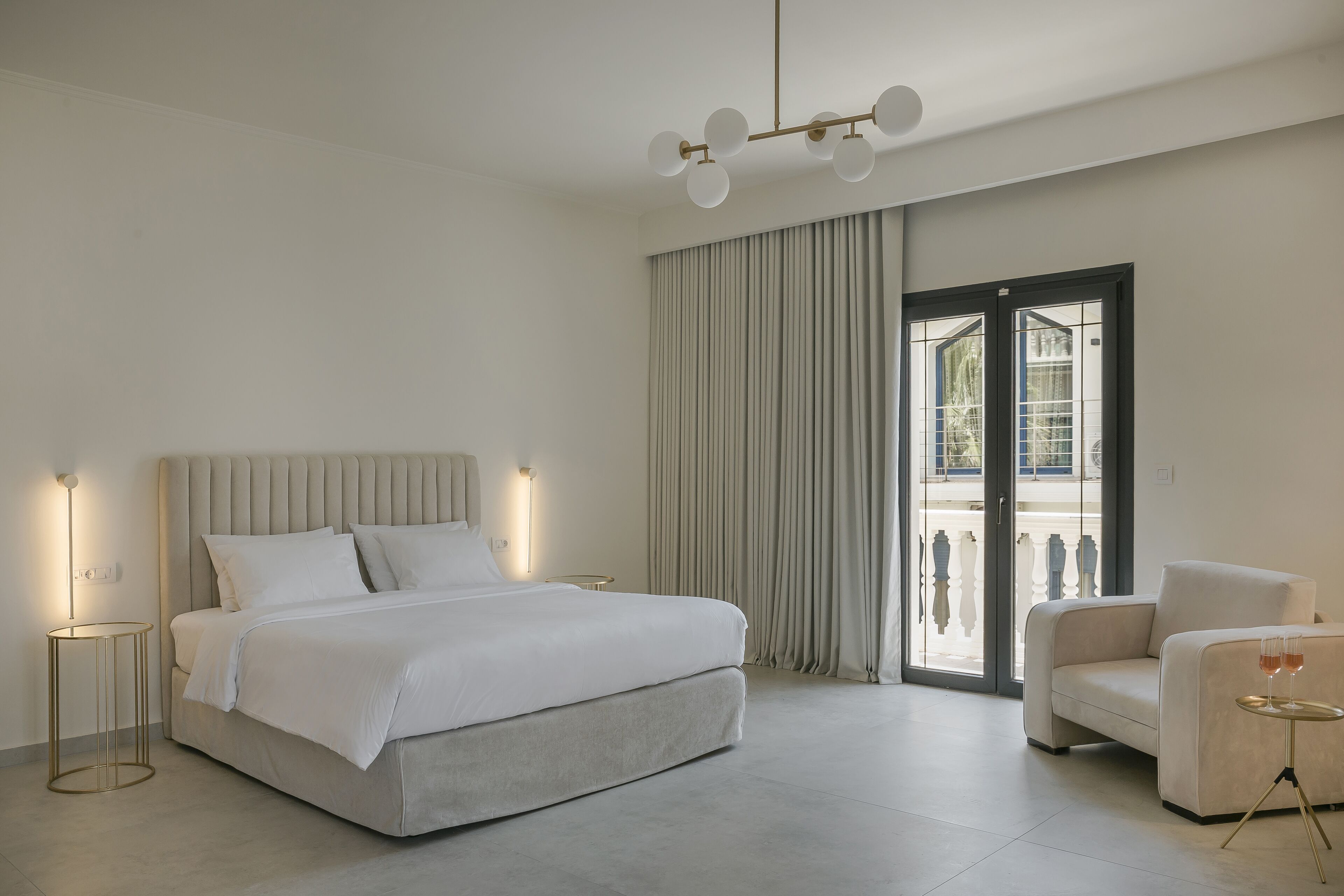 A modern bedroom with a large bed, a sofa, a small table with glasses, and a view through a glass door.