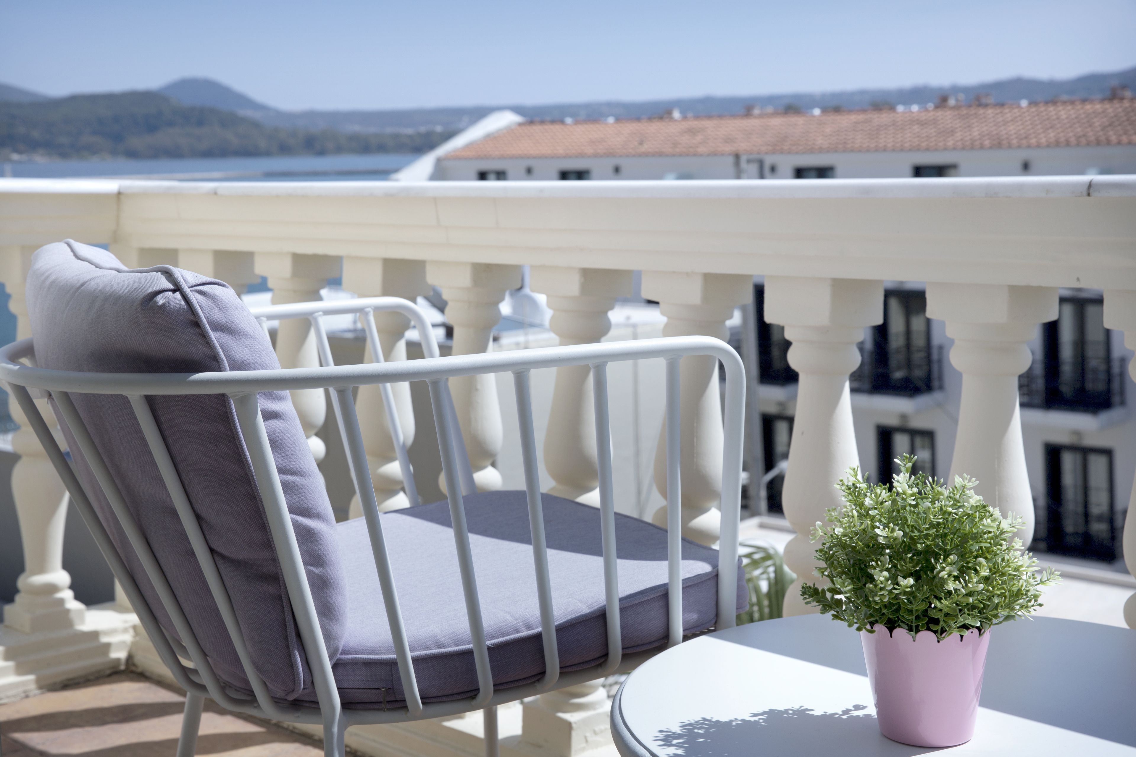 Un balcone con ringhiera in metallo bianco, una poltrona imbottita grigia e una piccola pianta in vaso.