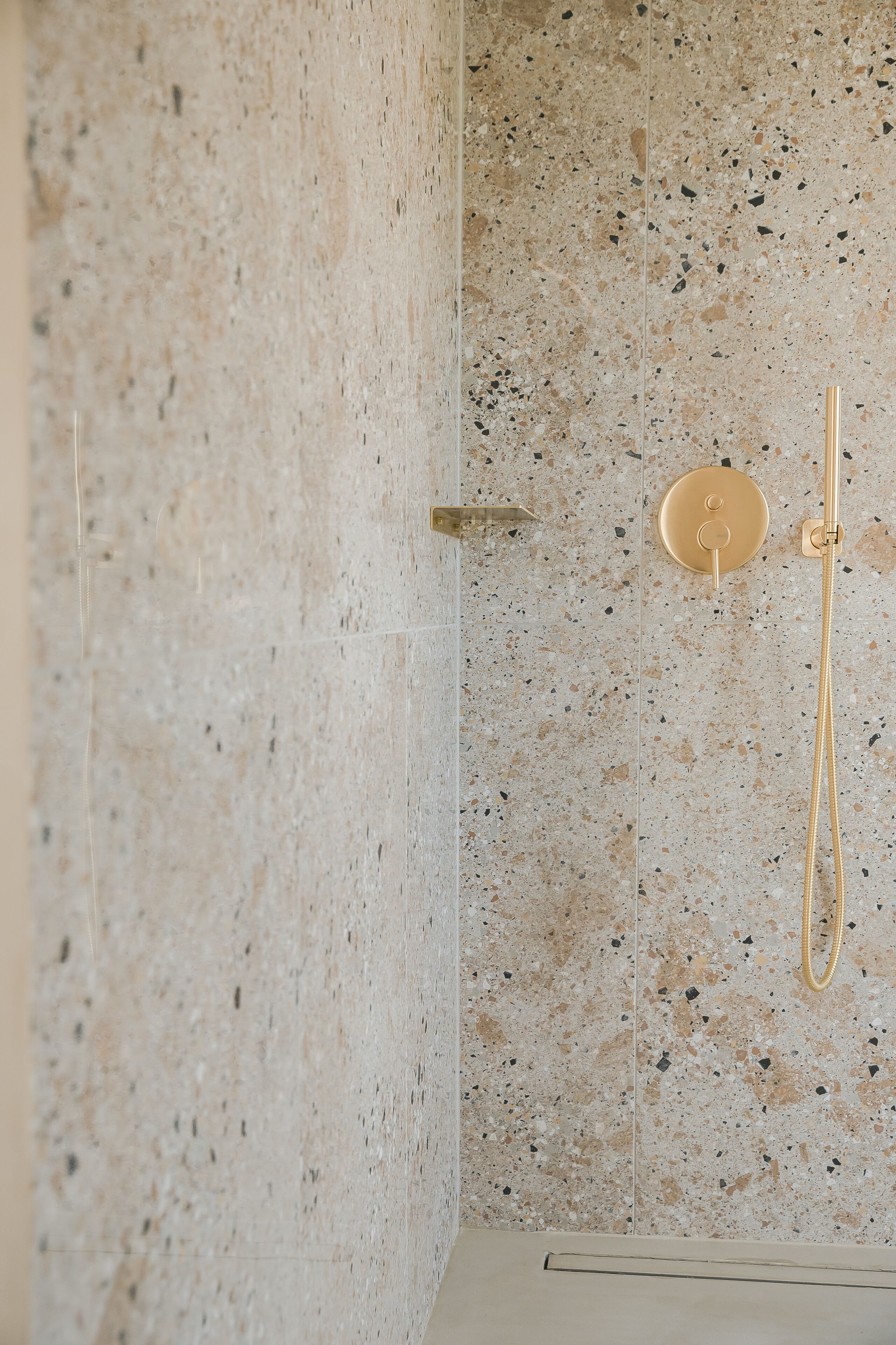 A shower with a gold shower head and hose, set against a speckled tile wall.