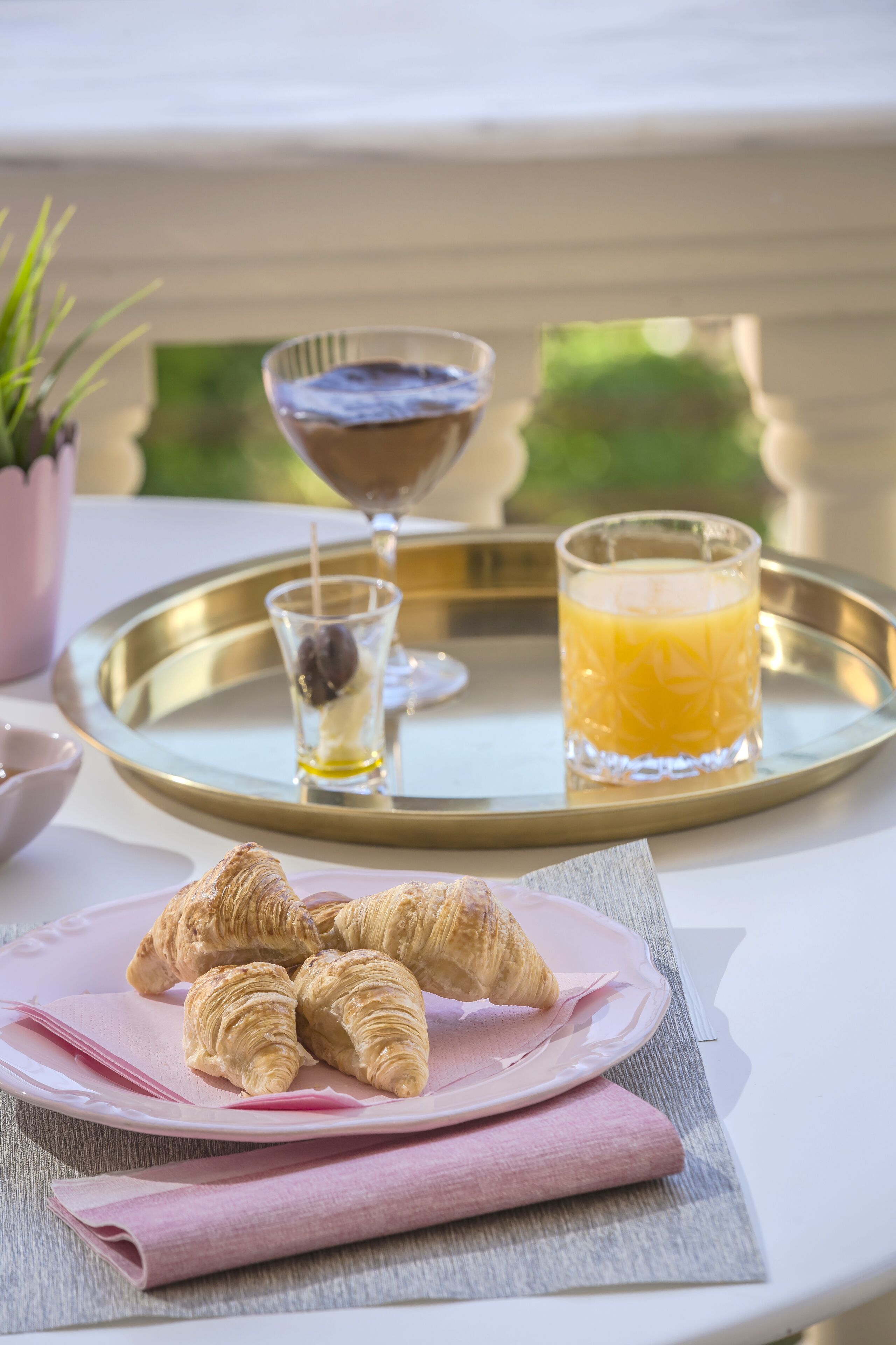 Una bandeja con croissants, un vaso de zumo de naranja y un chorrito de aceite de oliva sobre una mesa con una servilleta rosa.